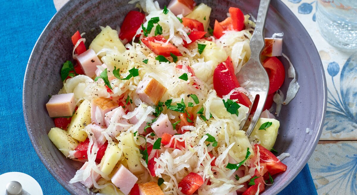 Sauerkrautsalat mit Kasseler in einer Schüssel auf einem blauen Untersetzer. Darunter eine Tischdecke mit blauen Blumen.