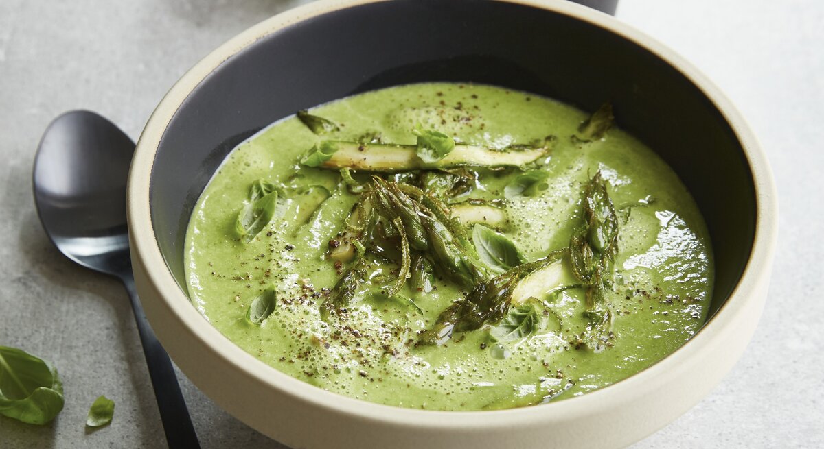 Eine graue Schale auf einem grauen Tisch mit der grünen Spargelcemesuppe. Alles von schräg oben fotografiert.
