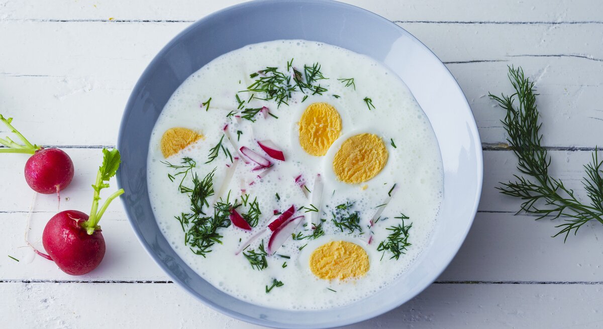 Kalte Sommersuppe mit Joghurt, Radieschen, Dill und hartgekochten Eiern in einer blauen Schale auf weißem Holztisch. Alles direkt von oben fotografiert.