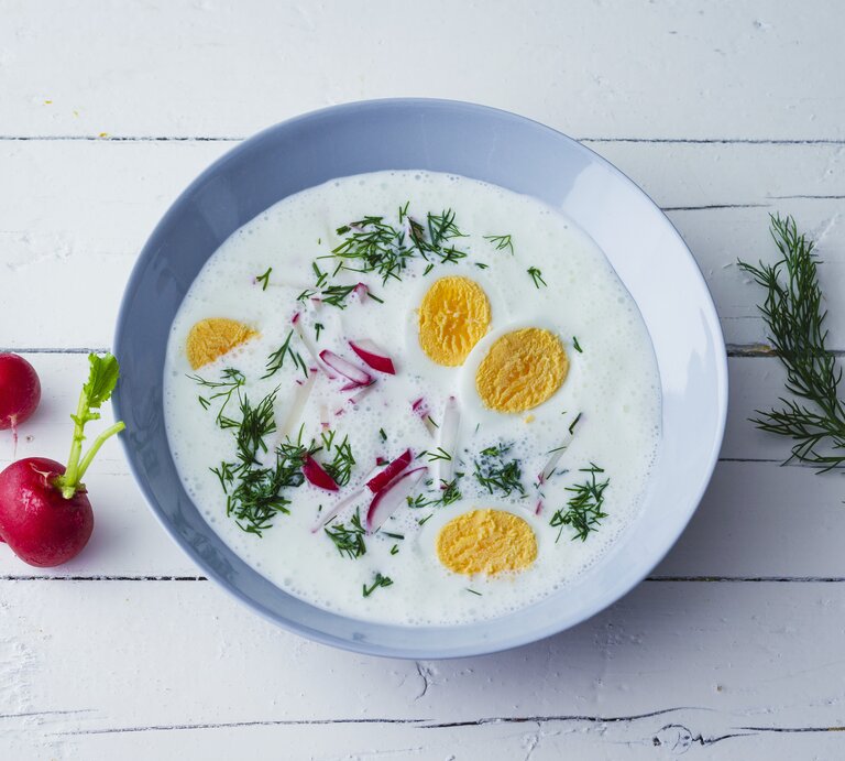 Kalte Sommersuppe mit Joghurt, Radieschen, Dill und hartgekochten Eiern in einer blauen Schale auf weißem Holztisch. Alles direkt von oben fotografiert. | © Stockfood
