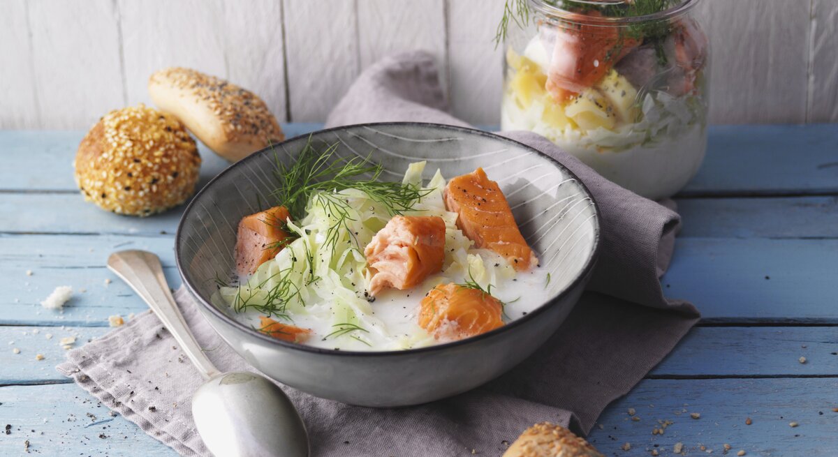 Eine graue Schüssel mit der Spitzkohlsuppe und Lachs auf einem hellblauem Holztisch von schräg oben fotografiert.