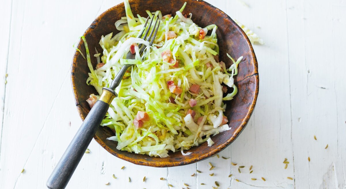 Draufsicht auf einen Bayrischen Krautsalat mit Speck in Schale. Das Ganze steht auf einem hellen Tisch. 