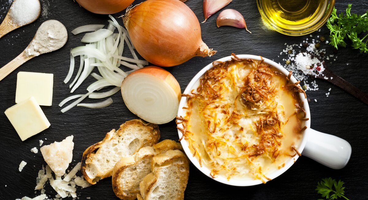  Das Bild zeigt eine appetitlich angerichtete französische Zwiebelsuppe in einem weißen Keramiktopf. Die Suppe ist mit geschmolzenem Käse überbacken, der goldbraun gratiniert ist. Um den Topf herum sind die Zutaten für die Suppe verteilt: frische Zwiebeln, geschnittene Zwiebelringe, Knoblauchzehen, ein Stück Butter, Olivenöl, eine Prise Salz und Pfeffer, sowie frisches Brot und geriebener Käse.