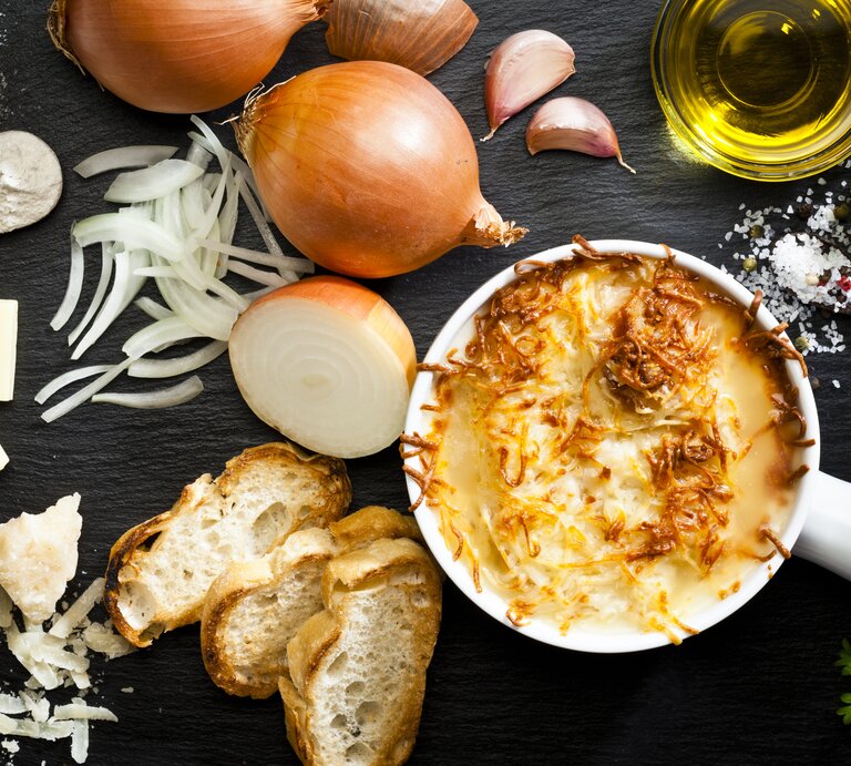  Das Bild zeigt eine appetitlich angerichtete französische Zwiebelsuppe in einem weißen Keramiktopf. Die Suppe ist mit geschmolzenem Käse überbacken, der goldbraun gratiniert ist. Um den Topf herum sind die Zutaten für die Suppe verteilt: frische Zwiebeln, geschnittene Zwiebelringe, Knoblauchzehen, ein Stück Butter, Olivenöl, eine Prise Salz und Pfeffer, sowie frisches Brot und geriebener Käse. | © iStock/fcafotodigital