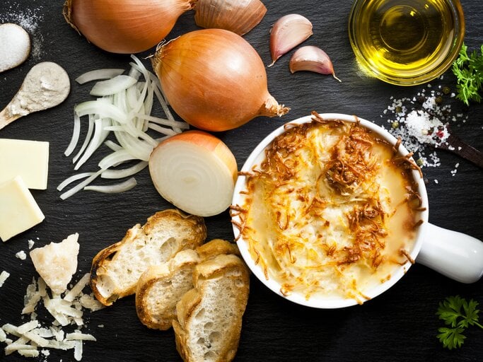  Das Bild zeigt eine appetitlich angerichtete französische Zwiebelsuppe in einem weißen Keramiktopf. Die Suppe ist mit geschmolzenem Käse überbacken, der goldbraun gratiniert ist. Um den Topf herum sind die Zutaten für die Suppe verteilt: frische Zwiebeln, geschnittene Zwiebelringe, Knoblauchzehen, ein Stück Butter, Olivenöl, eine Prise Salz und Pfeffer, sowie frisches Brot und geriebener Käse. | © iStock/fcafotodigital