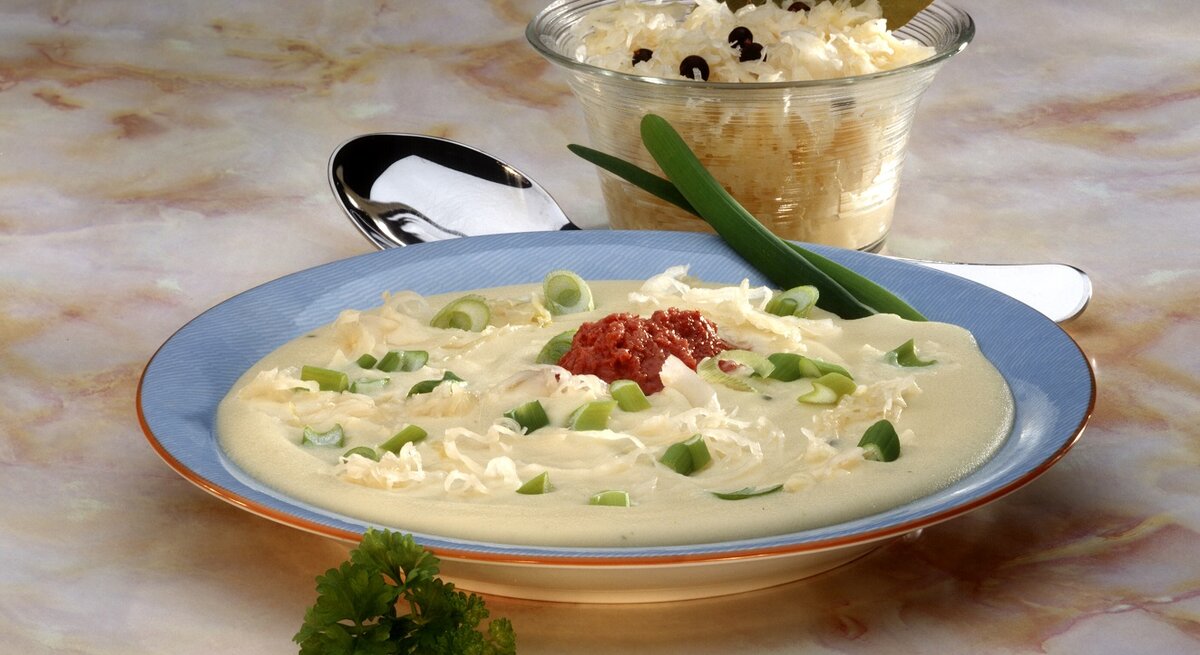 Eine Draufsicht auf einen Teller mit cremiger Suppe, garniert mit frischen grünen Zwiebelringen, geriebenem Käse und einem Klecks roter Soße in der Mitte. Die Suppe wird in einem blauen Suppenteller mit einem orangefarbenen Rand serviert. Im Hintergrund befindet sich eine Glasschale mit weiterem geriebenem Käse, verziert mit Lorbeerblättern und Pfefferkörnern. Die Szene liegt auf einer Marmor-Tischoberfläche und wird von einem silbernen Löffel und einem frischen Petersilienzweig ergänzt. 
