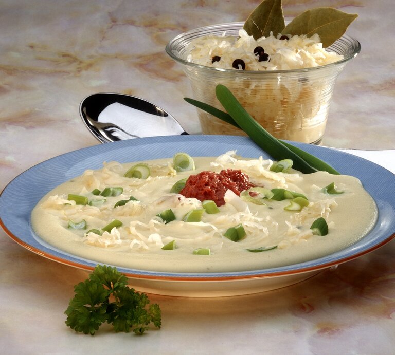 Eine Draufsicht auf einen Teller mit cremiger Suppe, garniert mit frischen grünen Zwiebelringen, geriebenem Käse und einem Klecks roter Soße in der Mitte. Die Suppe wird in einem blauen Suppenteller mit einem orangefarbenen Rand serviert. Im Hintergrund befindet sich eine Glasschale mit weiterem geriebenem Käse, verziert mit Lorbeerblättern und Pfefferkörnern. Die Szene liegt auf einer Marmor-Tischoberfläche und wird von einem silbernen Löffel und einem frischen Petersilienzweig ergänzt.  | © StockFood/Bohle, Gaby