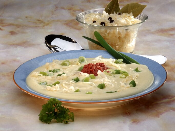 Eine Draufsicht auf einen Teller mit cremiger Suppe, garniert mit frischen grünen Zwiebelringen, geriebenem Käse und einem Klecks roter Soße in der Mitte. Die Suppe wird in einem blauen Suppenteller mit einem orangefarbenen Rand serviert. Im Hintergrund befindet sich eine Glasschale mit weiterem geriebenem Käse, verziert mit Lorbeerblättern und Pfefferkörnern. Die Szene liegt auf einer Marmor-Tischoberfläche und wird von einem silbernen Löffel und einem frischen Petersilienzweig ergänzt.  | © StockFood/Bohle, Gaby