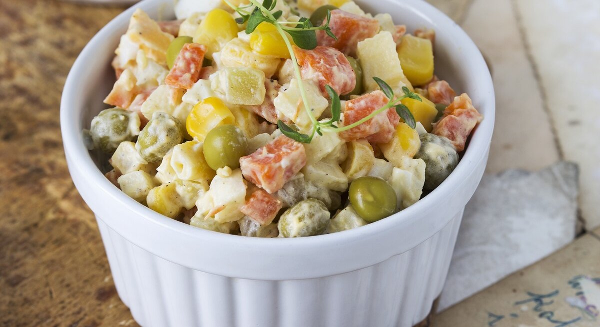 Eine kleine weiße Schale gefüllt mit einem Kartoffelsalat mit Mais, Ei, Erbsen, Karotten und Mayonnaise.