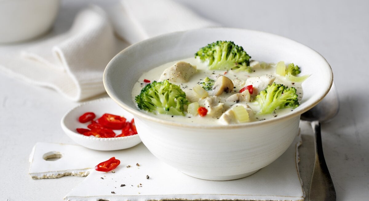 Eine weiße Schale mit der Hähnchen-Brokkoli-Suppe auf einem weißen Tisch von der Seite fotografiert.