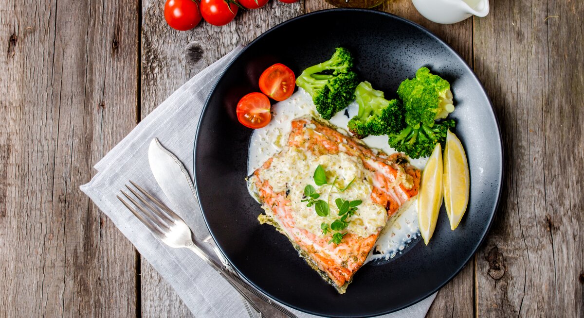 Draufsicht auf einen schwarzen Teller mit einem Stück Lachs und einer cremigen Weißweinsoße. Daneben Zitronenschnitze, Tomaten und Brokkoli. Das ganze steht auf einem dekorierten Holztisch. 