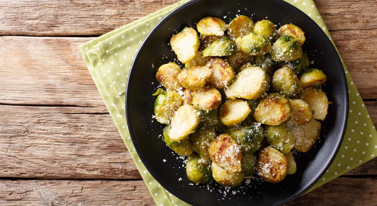Draufsicht auf gebratenen Rosenkohl mit Parmesan auf schwarzem Teller. Dieser steht auf Holztisch.