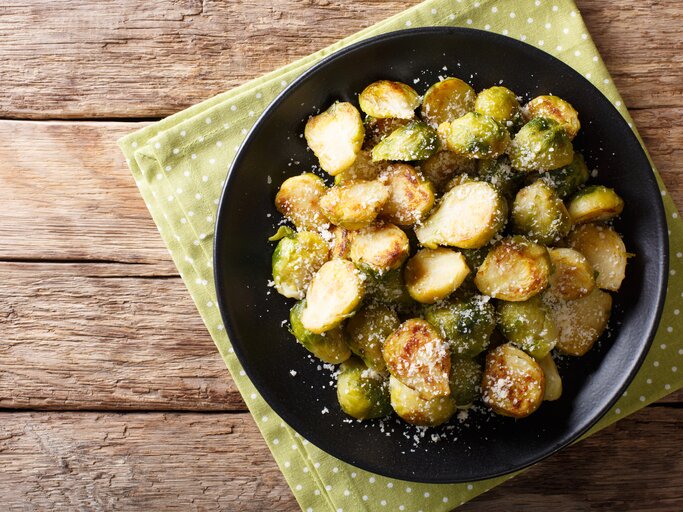 Draufsicht auf gebratenen Rosenkohl mit Parmesan auf schwarzem Teller. Dieser steht auf Holztisch. | © iStock/ALLEKO
