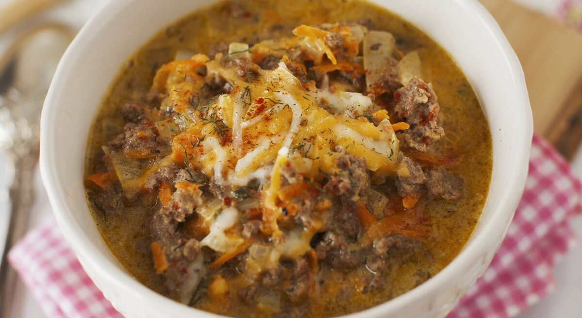Eine weiße Schüssel mit herzhafter, cremiger Suppe aus Hackfleisch und Gemüse, garniert mit geschmolzenem Käse auf kariertem Tuch.