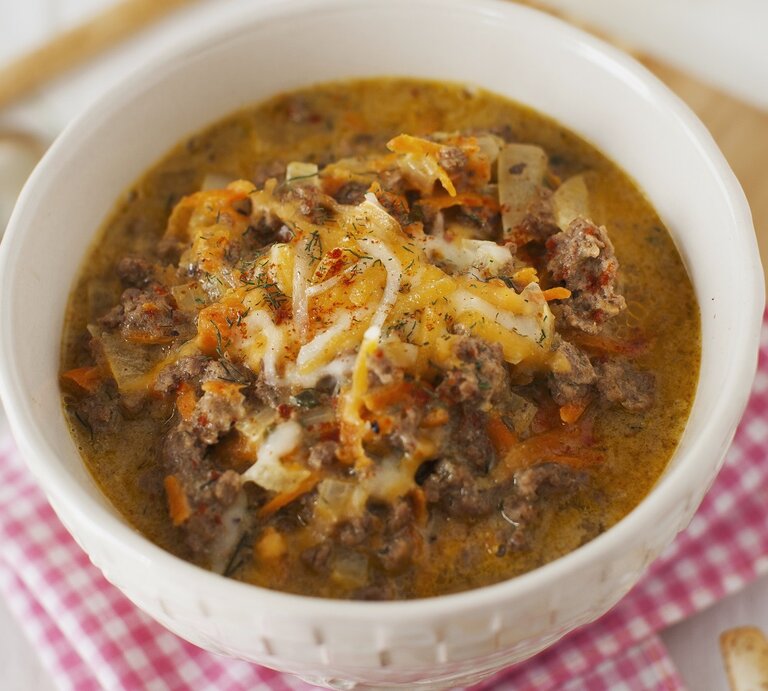 Eine weiße Schüssel mit herzhafter, cremiger Suppe aus Hackfleisch und Gemüse, garniert mit geschmolzenem Käse auf kariertem Tuch. | © StockFood/Strokin, Yelena