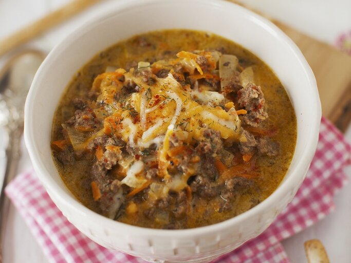 Eine weiße Schüssel mit herzhafter, cremiger Suppe aus Hackfleisch und Gemüse, garniert mit geschmolzenem Käse auf kariertem Tuch. | © StockFood/Strokin, Yelena