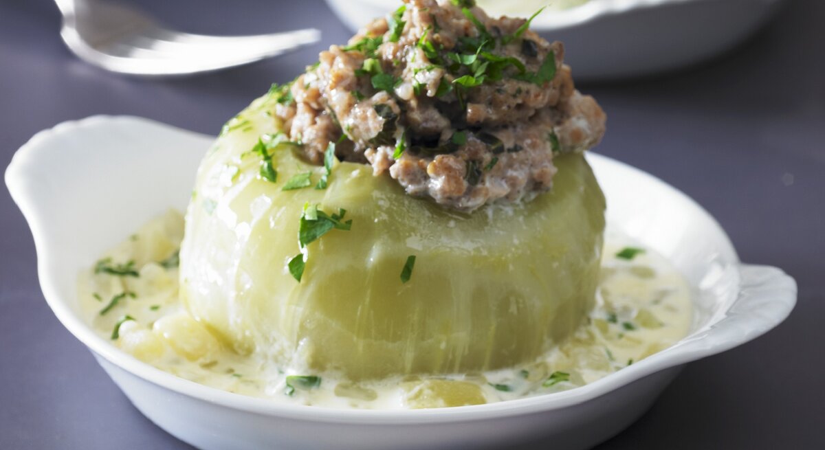 Gefüllter Kohlrabi mit Hackfleisch in einem tiefen weißem Teller auf grauem Untergrund. Im Hintergrund ist unscharf eine zweite Portion im Teller zu sehen.