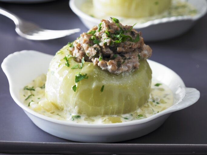 Gefüllter Kohlrabi mit Hackfleisch in einem tiefen weißem Teller auf grauem Untergrund. Im Hintergrund ist unscharf eine zweite Portion im Teller zu sehen. | © Stockfood/Walter Cimbal