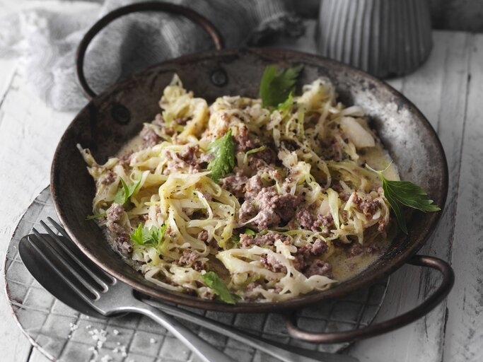 Eine schwarze Pfanne mit dem Gericht auf einem hellgrauen Tisch mit dunkler Dekoration. Alles von schräg oben fotografiert. | © StockFood / Gräfe & Unzer Verlag / Kramp + Gölling