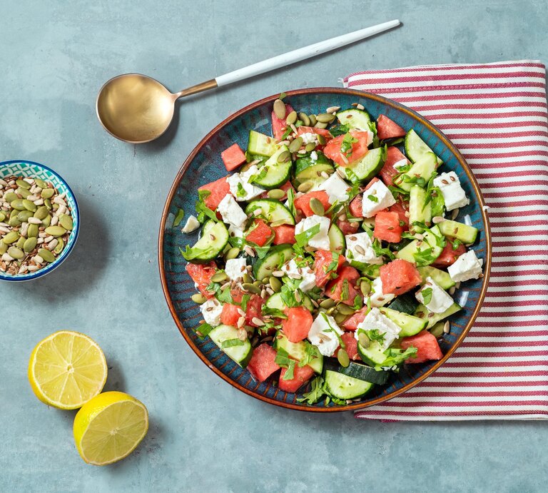 Gurkensalat mit Wassermelone und Feta auf blauem Teller, rot-weißem Tuch und hellblauem Untergrund. Im BIld eine Lössel, eine Schale mit Kernen und eine geteilte Zitrone. Draufsicht. | © Getty Images / Carlo A