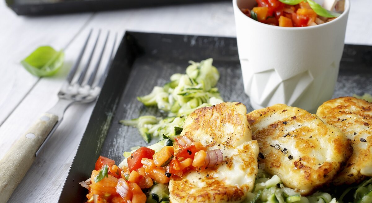 Draufsicht, schräg: Auf einem Untergrund aus hellem Holz liegt ein dunkles Tablett aus Holz. Darauf ist gebratener Halloumi auf Zucchinisalat.