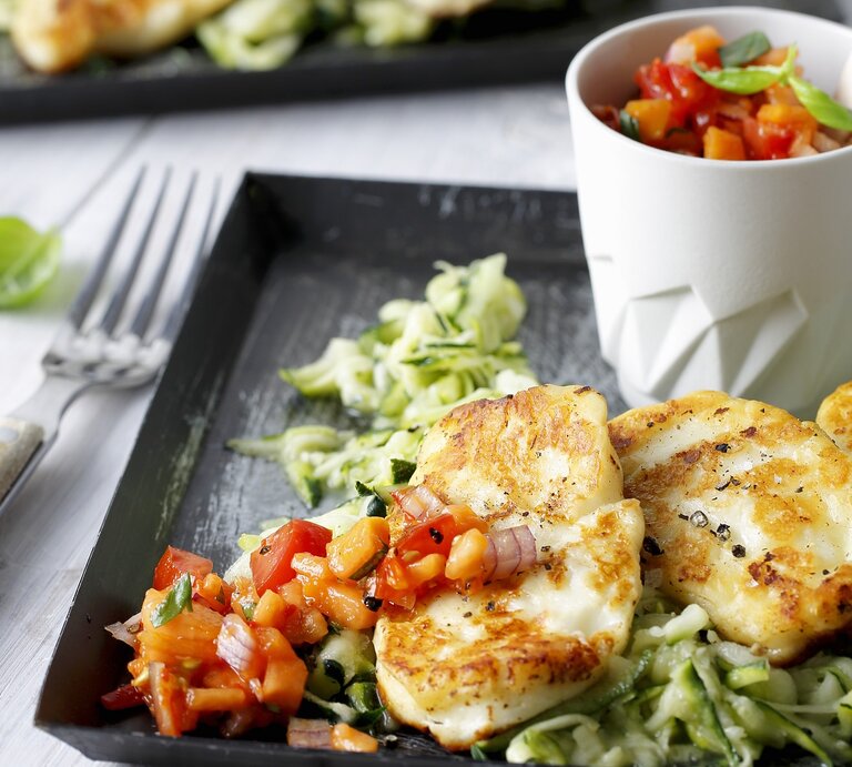 Draufsicht, schräg: Auf einem Untergrund aus hellem Holz liegt ein dunkles Tablett aus Holz. Darauf ist gebratener Halloumi auf Zucchinisalat. | © Stockfood/Gräfe & Unzer Verlag / mona binner PHOTOGRAPHIE