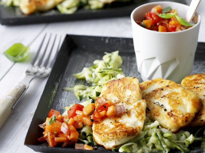 Draufsicht, schräg: Auf einem Untergrund aus hellem Holz liegt ein dunkles Tablett aus Holz. Darauf ist gebratener Halloumi auf Zucchinisalat. | © Stockfood/Gräfe & Unzer Verlag / mona binner PHOTOGRAPHIE