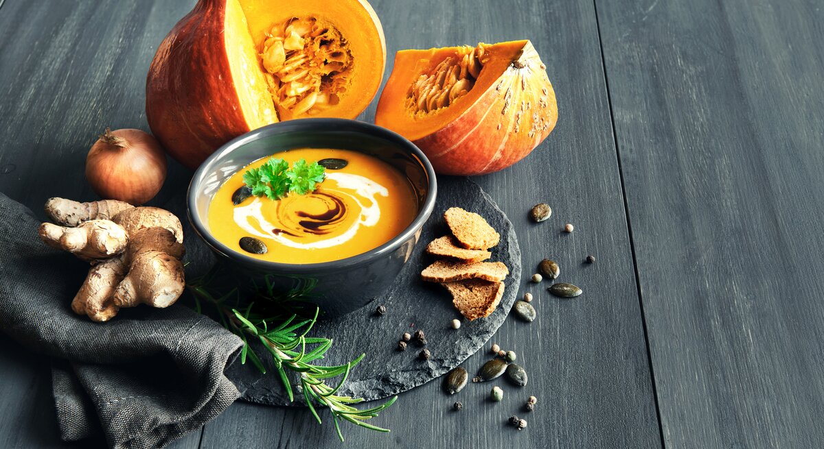 Schwarze Schale gefüllt mit Kürbsisuppe, garniert mit Creme und Kürbiskernen. Daneben liegt ein angeschnittener Kürbis. 