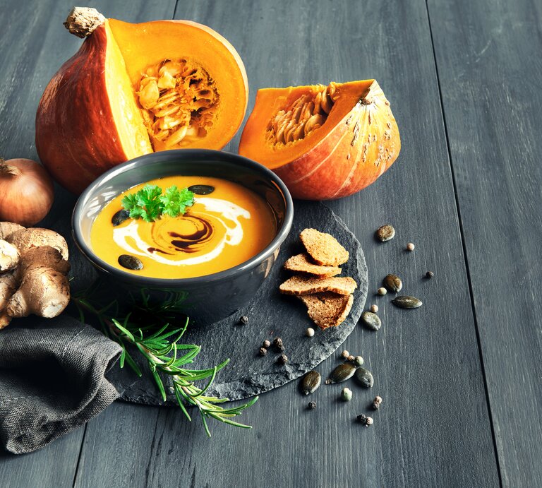 Schwarze Schale gefüllt mit Kürbsisuppe, garniert mit Creme und Kürbiskernen. Daneben liegt ein angeschnittener Kürbis.  | © iStock/anyaivanova