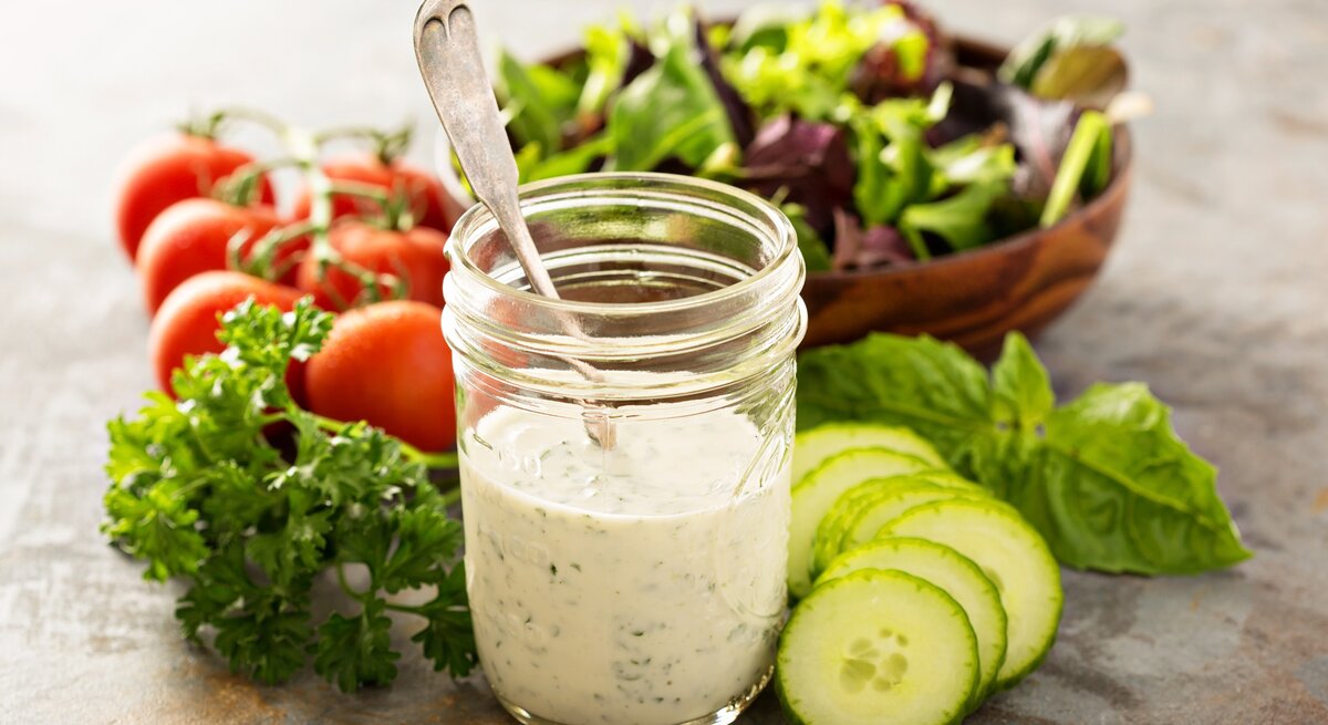 Frisch angerührtes Joghurtdressing mit frischen Kräutern im Glas. Daneben liegen knackige Salatzutaten.
