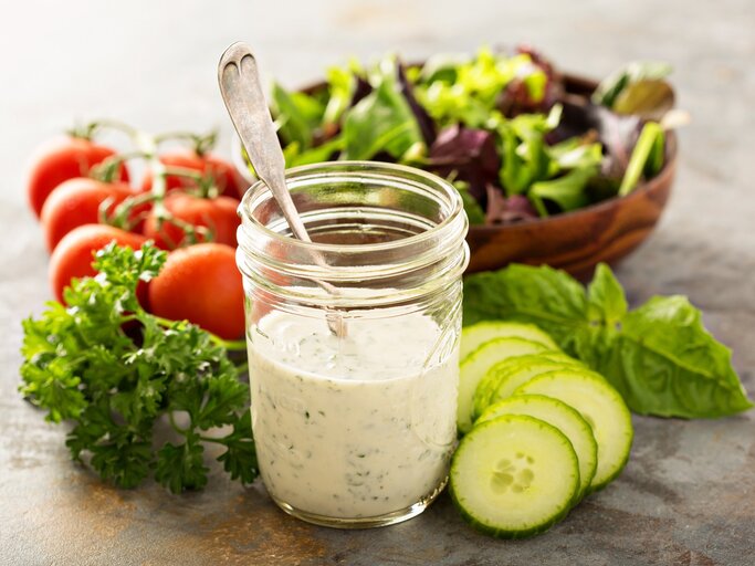 Frisch angerührtes Joghurtdressing mit frischen Kräutern im Glas. Daneben liegen knackige Salatzutaten. | © iStock/VeselovaElena