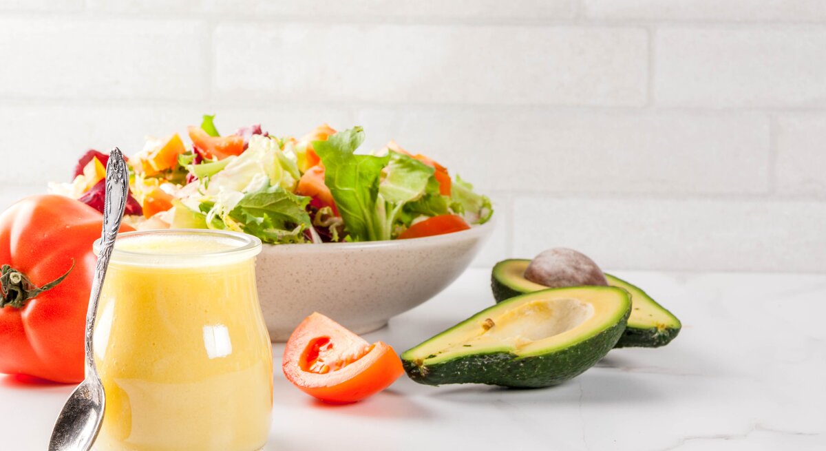 Ein Glas mit Honig-Senf-Dressing steht auf einem weißen Tisch, dahinter eine schale Salat. 