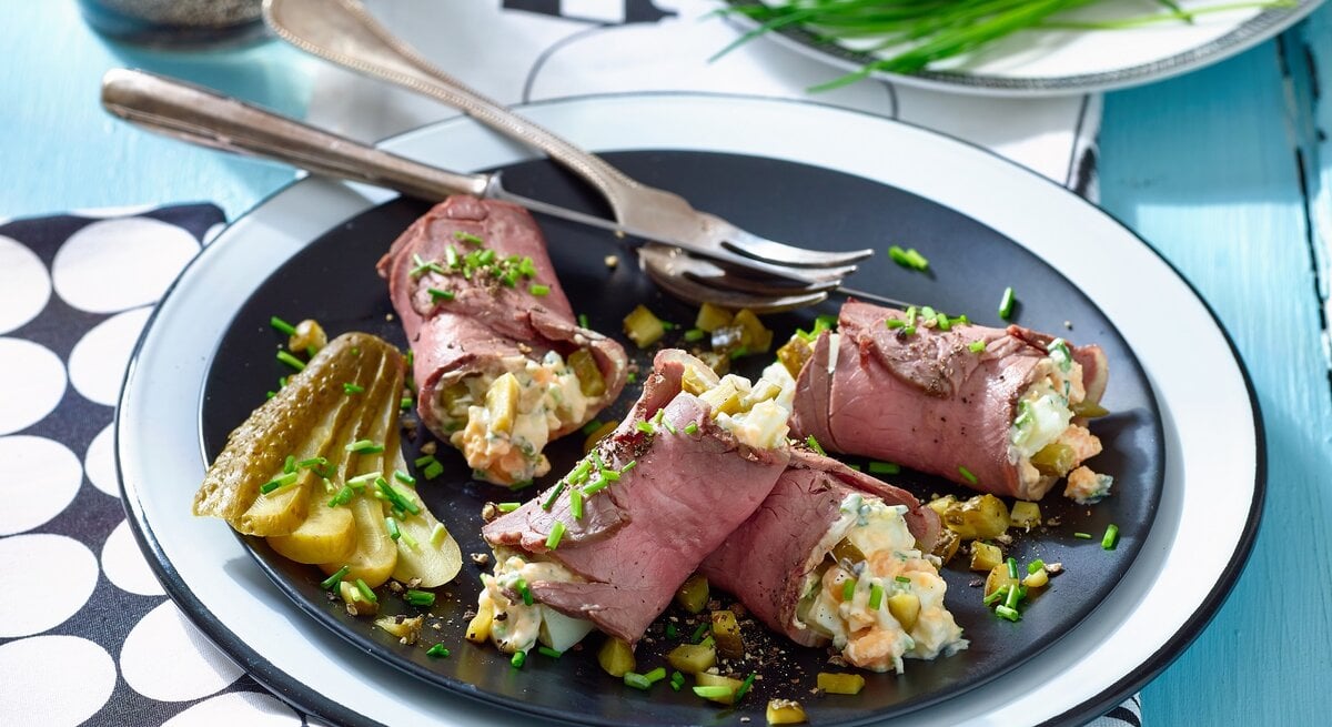 Roastbeef mit Eiersalat auf einem Teller auf einem schwarz-weiß-gemusterten Tischtuch. Hinten auf den Foto ein Teller mit Essiggurken und Schnittlauch.
