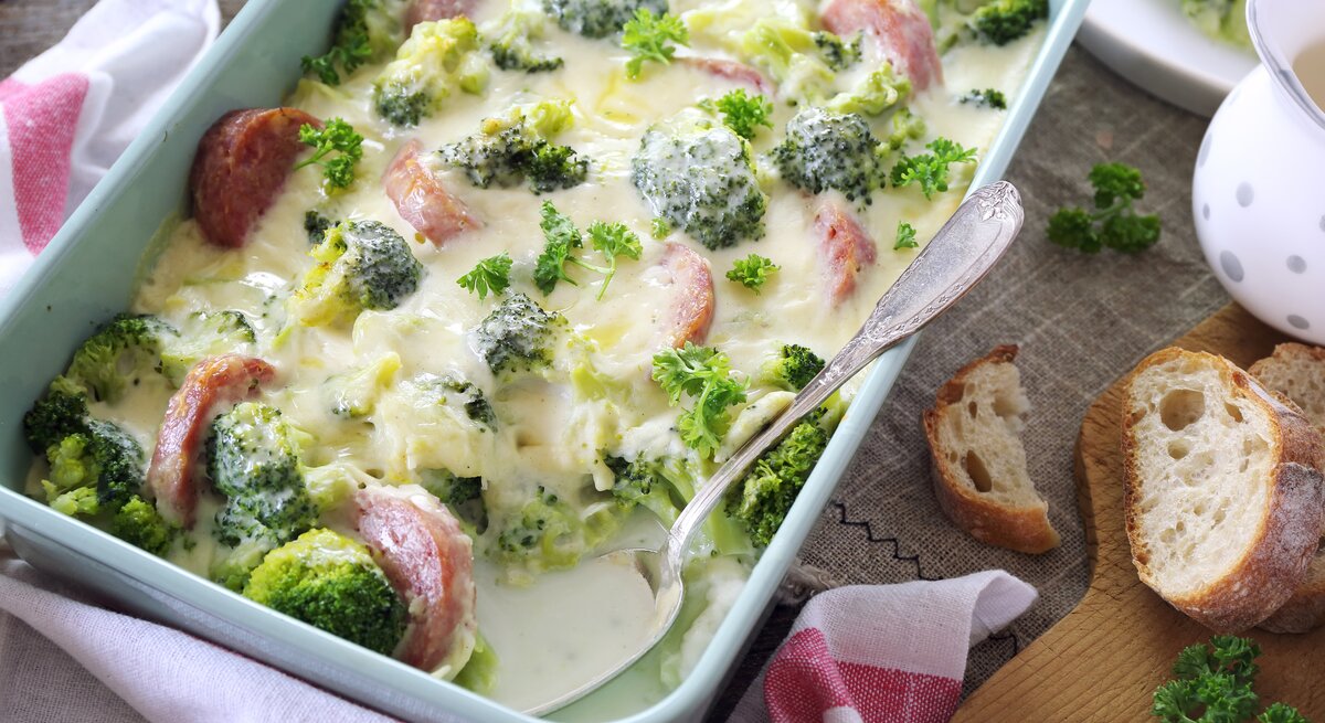 Dieses Bild zeigt eine Auflaufform mit einem cremigen Brokkoli- und Käsegericht mit Scheiben von geräucherter Wurst, das appetitlich aussieht und zum Essen bereit ist. Das Essen ist wahrscheinlich warm, da es aussieht, als wäre es gerade aus dem Ofen gekommen. Auf dem Tisch neben der Auflaufform befinden sich eine Scheibe Brot, ein Bund Petersilie und ein Stückchen Brot, was darauf hindeutet, dass das Gericht möglicherweise mit Brot serviert wird, um die cremige Sauce aufzunehmen. 