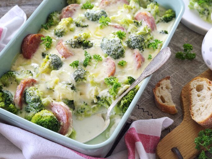 Dieses Bild zeigt eine Auflaufform mit einem cremigen Brokkoli- und Käsegericht mit Scheiben von geräucherter Wurst, das appetitlich aussieht und zum Essen bereit ist. Das Essen ist wahrscheinlich warm, da es aussieht, als wäre es gerade aus dem Ofen gekommen. Auf dem Tisch neben der Auflaufform befinden sich eine Scheibe Brot, ein Bund Petersilie und ein Stückchen Brot, was darauf hindeutet, dass das Gericht möglicherweise mit Brot serviert wird, um die cremige Sauce aufzunehmen.  | © iStock.com/photosimysia