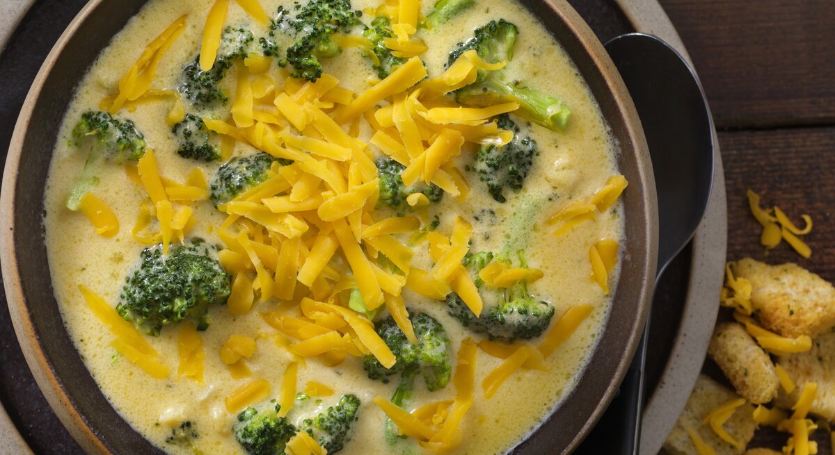 Das Bild zeigt eine Schale mit Brokkoli-Käse-Suppe. Die Suppe hat eine cremige Konsistenz und ist mit viel geriebenem Cheddar-Käse bestreut, was für eine reiche und ansprechende Textur sorgt. Die leuchtend grünen Brokkoliröschen schwimmen in der gelblichen Suppe und bieten einen schönen Farbkontrast. Am Rand der Schale sind einige Croutons zu sehen, die als Beilage dienen könnten. Der dunkle Holzhintergrund verleiht dem Bild Wärme und betont die appetitliche Präsentation der Speise.
