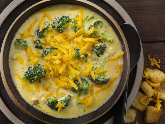 Das Bild zeigt eine Schale mit Brokkoli-Käse-Suppe. Die Suppe hat eine cremige Konsistenz und ist mit viel geriebenem Cheddar-Käse bestreut, was für eine reiche und ansprechende Textur sorgt. Die leuchtend grünen Brokkoliröschen schwimmen in der gelblichen Suppe und bieten einen schönen Farbkontrast. Am Rand der Schale sind einige Croutons zu sehen, die als Beilage dienen könnten. Der dunkle Holzhintergrund verleiht dem Bild Wärme und betont die appetitliche Präsentation der Speise. | © iStock.com/LauriPatterson