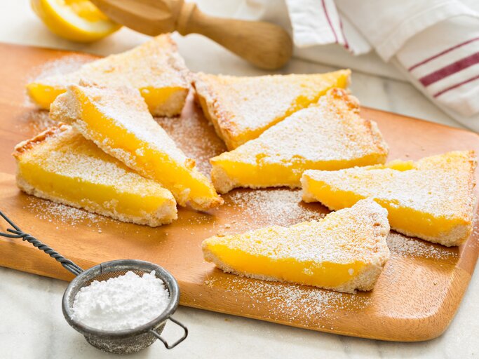 Zitronenkuchenstücke auf einem Holzbrett mit Puderzucker bestäubt, daneben ein Sieb und eine angeschnittene Zitrone im Hintergrund. | © iStock/DebbiSmirnoff