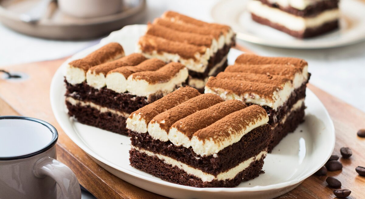 Ein weißer Teller mit drei Stücken der Tiramisu-Torte mit Mascarponecreme auf einem dunklen Holztisch.