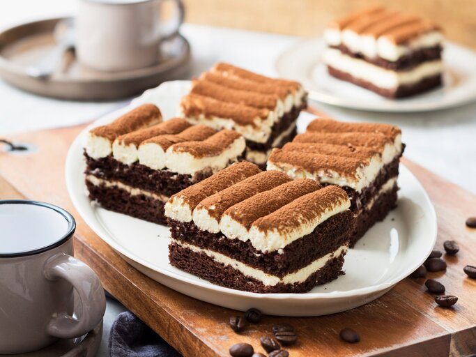 Ein weißer Teller mit drei Stücken der Tiramisu-Torte mit Mascarponecreme auf einem dunklen Holztisch. | © StockFood / ZS Verlag / Speck, Sabrina