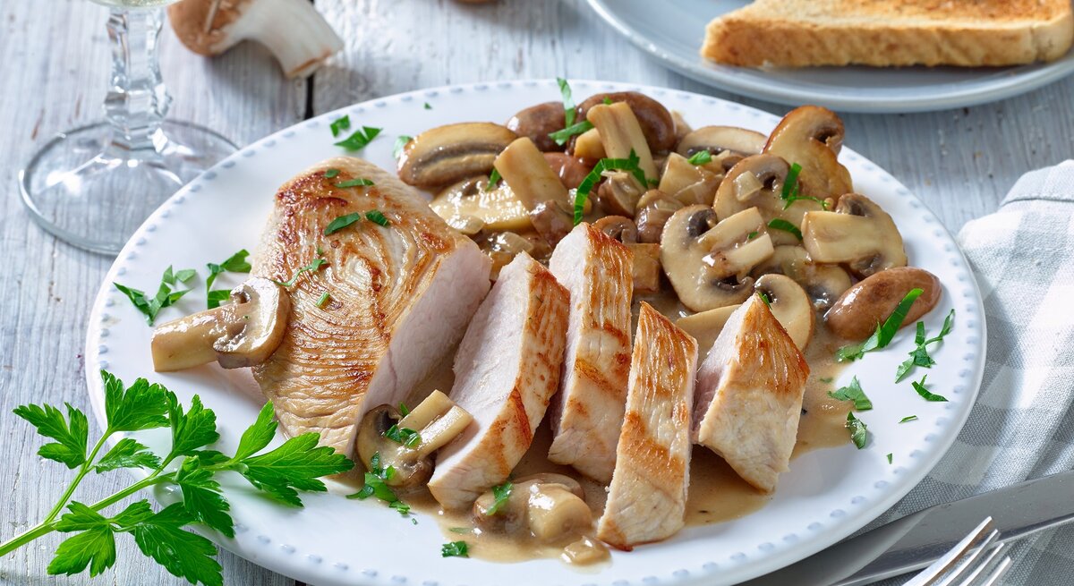 Putenschnitzel mit Champignons auf einem weißen Teller. Rechts Besteck und links ein Weinglas. Der Hintergrund ist hell und Champignons darauf verteilt.