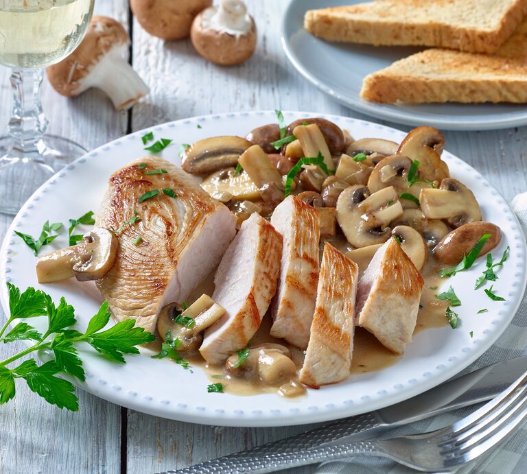 Putenschnitzel mit Champignons auf einem weißen Teller. Rechts Besteck und links ein Weinglas. Der Hintergrund ist hell und Champignons darauf verteilt. | © Matthias Liebich