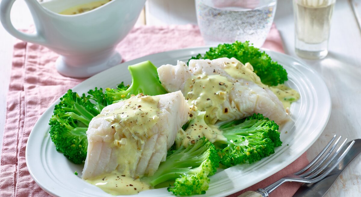 Zanderfilet mit Brokkoli auf einem runden Teller aus Porzellan / Messer und Gabel / ein Behältnis aus Porzellan mit Sauce / Ein Glas mit Sprudelwasser 