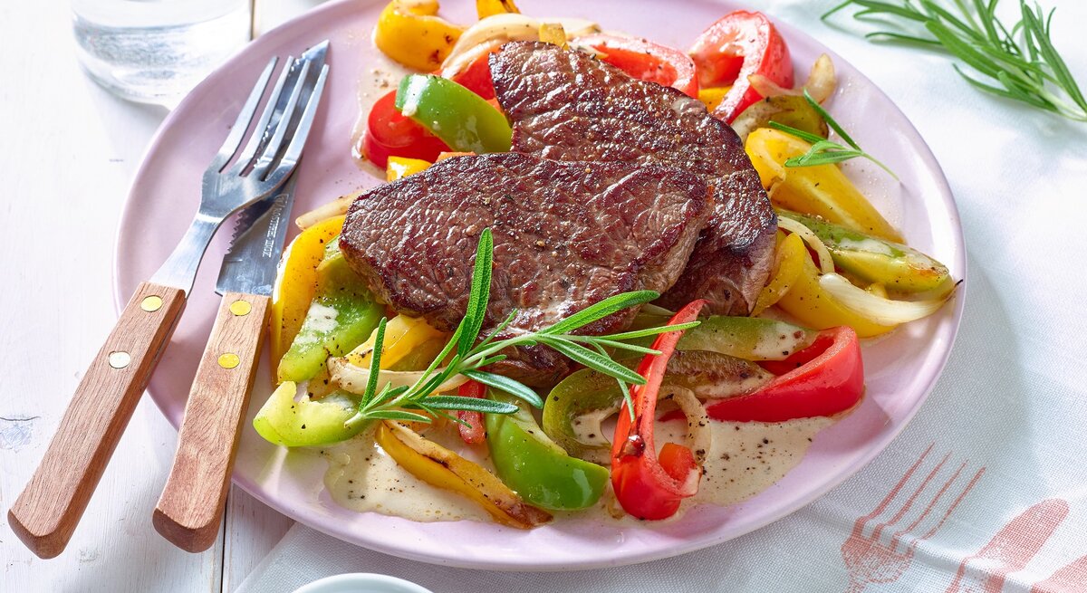 Rindersteak mit Paprikagemüse auf einem rosa Teller, links das Besteck. Der Hintergrund ist weiß.