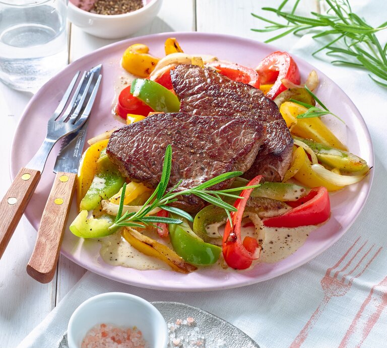 Rindersteak mit Paprikagemüse auf einem rosa Teller, links das Besteck. Der Hintergrund ist weiß. | © Matthias Liebich
