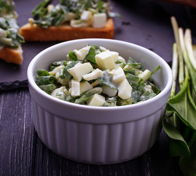 Bärlauch-Salat mit Käse in einer weißen Schale, angerichtet auf einem dunklen Holztisch.  | © iStock/ yulka3ice