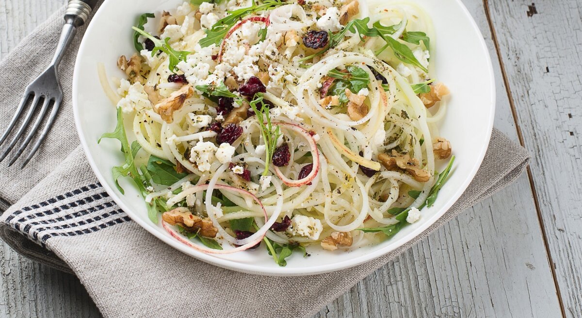 Draufsicht: Ein runder Teller mit einem Kohlrabi-Apfel-Salat mit Käse und Walnüssen auf einem hellen Untergrund aus Holz.