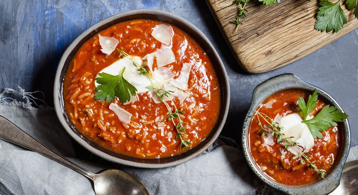 Tomatensuppe mit Reis, Hackfleisch und Parmesanspänen in einer grauen Schale auf einem dunkelblauen Steintisch. Alles direkt von oben fotografiert.