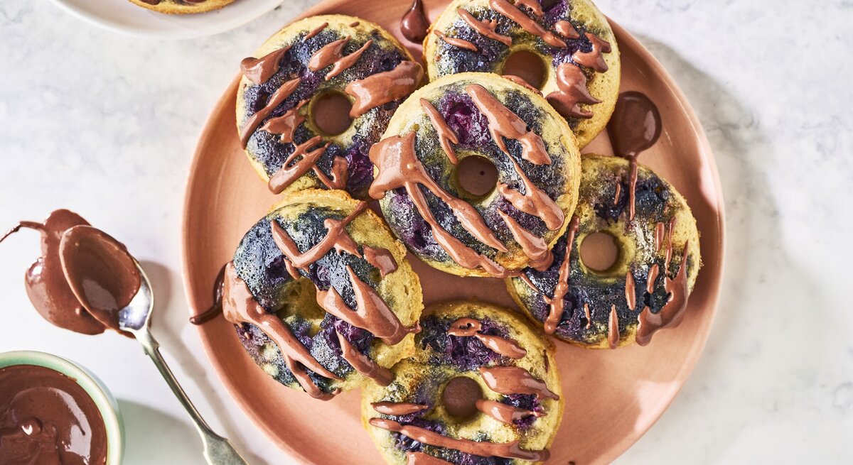 Draufsicht: Weißer Marmoruntergrund auf dem in der Bildmitte ein Tonteller mit sechs Donuts steht. Links daneben steht ein kleines Schüsselchen mit Mandel-Schokoladen-Topping und einem Löffel. In der oberen linken Bildecke steht ein weiterer, weißer Teller mit einem einzelnen Donut drauf.