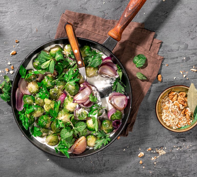 Draufsicht auf eine runde Pfanne mit hölzernem Stiel. In der pfanne ist gebratener Rosenkohl und Joghurtsoße.  | © iStock/KucherAV